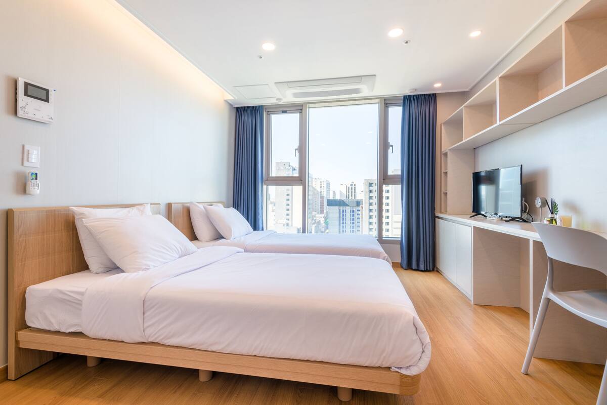 A modern hotel room with two beds, a desk, and a large window with a city view.