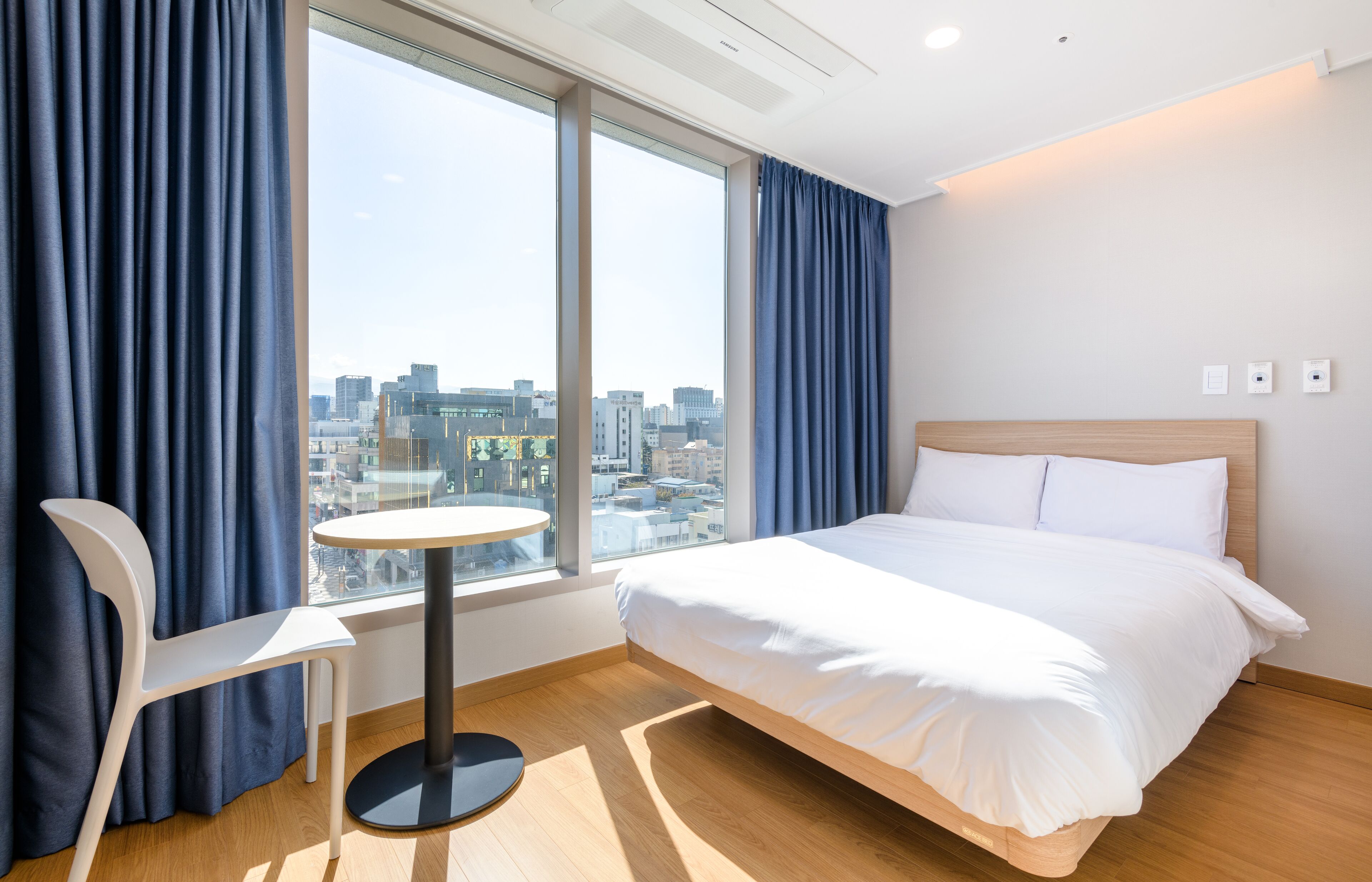 A hotel room with a bed, a round table, a chair, and a large window offering a city view.