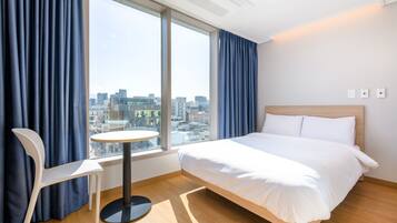 A hotel room with a bed, a round table, a chair, and a large window offering a city view.
