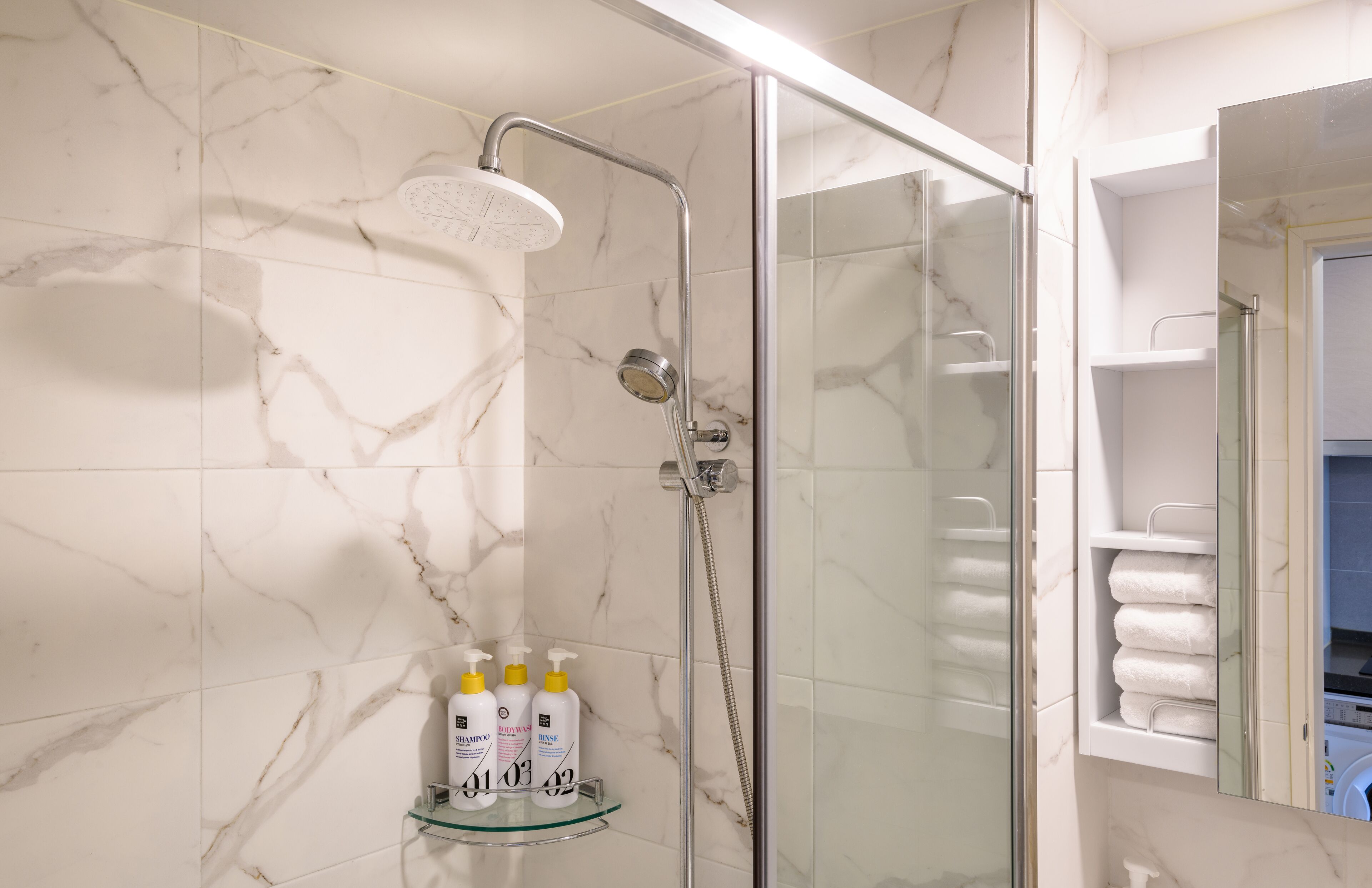 A modern bathroom with a glass-enclosed shower, marble walls, and a glass shelf holding toiletries.