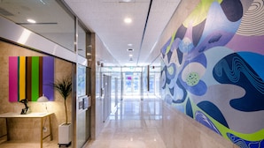Un pasillo moderno con entrada por una puerta de cristal, un mural abstracto y colorido a la derecha, y un mostrador de recepción con una lámpara y una estatua a la izquierda.