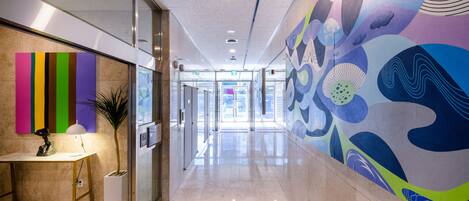 A modern hallway with a glass door entrance, a colorful abstract mural on the right, and a reception desk with a lamp and a statue on the left.