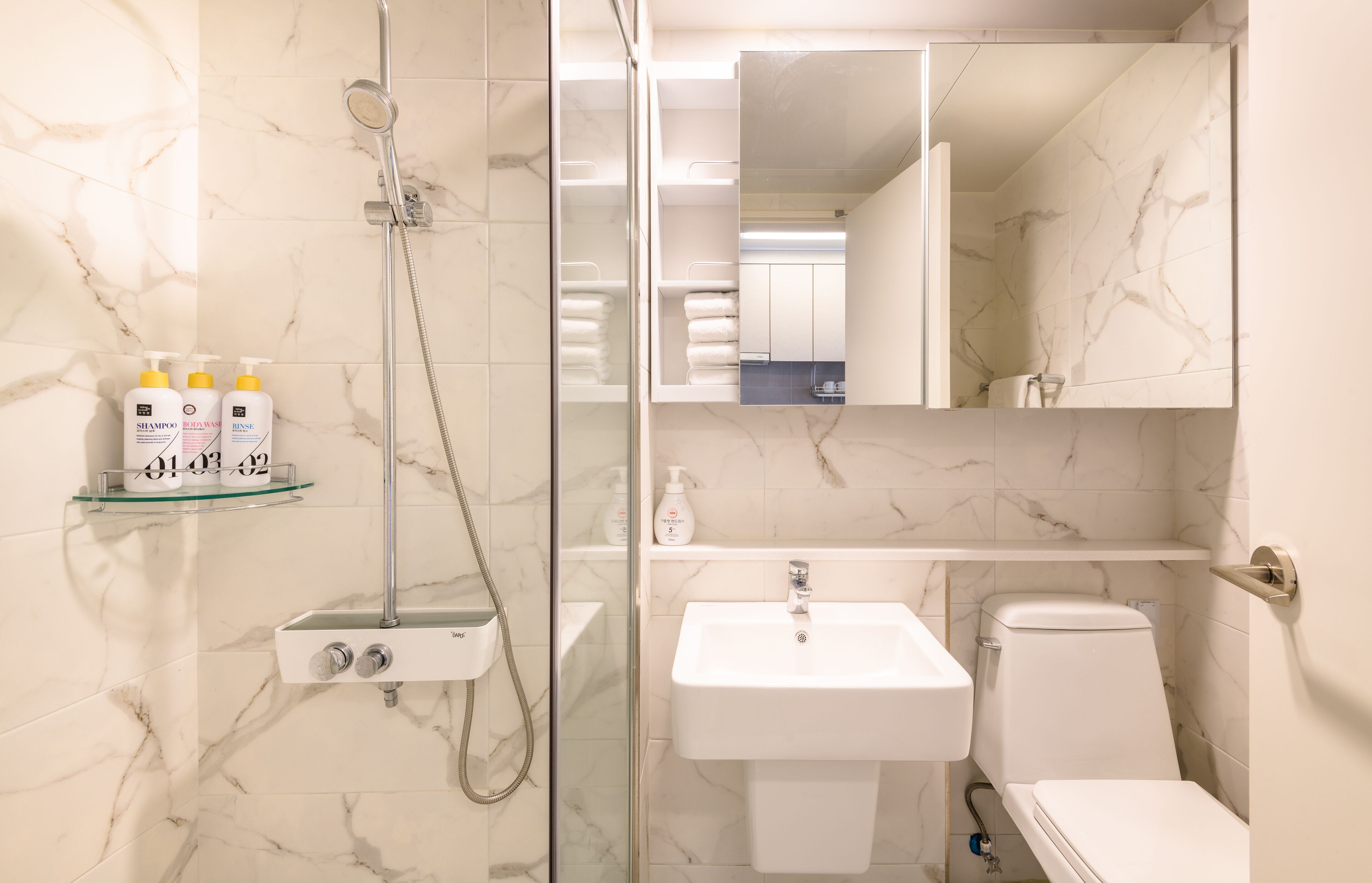 A bathroom with a glass-enclosed shower, a white sink, and a toilet. There are toiletries on a shelf.