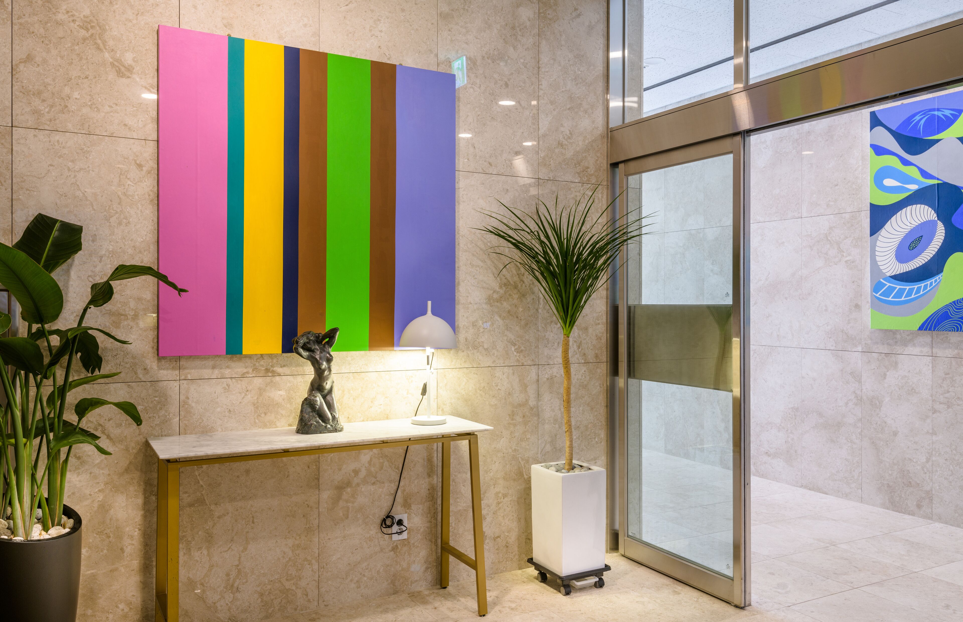 Un vestíbulo con piso de mármol, una obra de arte vertical y colorida, un escritorio con lámpara, una planta en maceta y una puerta de vidrio con vista al exterior.