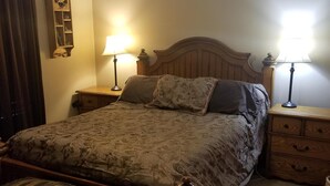 A bedroom with a wooden bed, two bedside tables with lamps, a ceiling fan, a wooden chest, and a wall-mounted shelf with decorative items. - American Pines Lodge, 5 miles from Mt. Rushmore (Keystone)