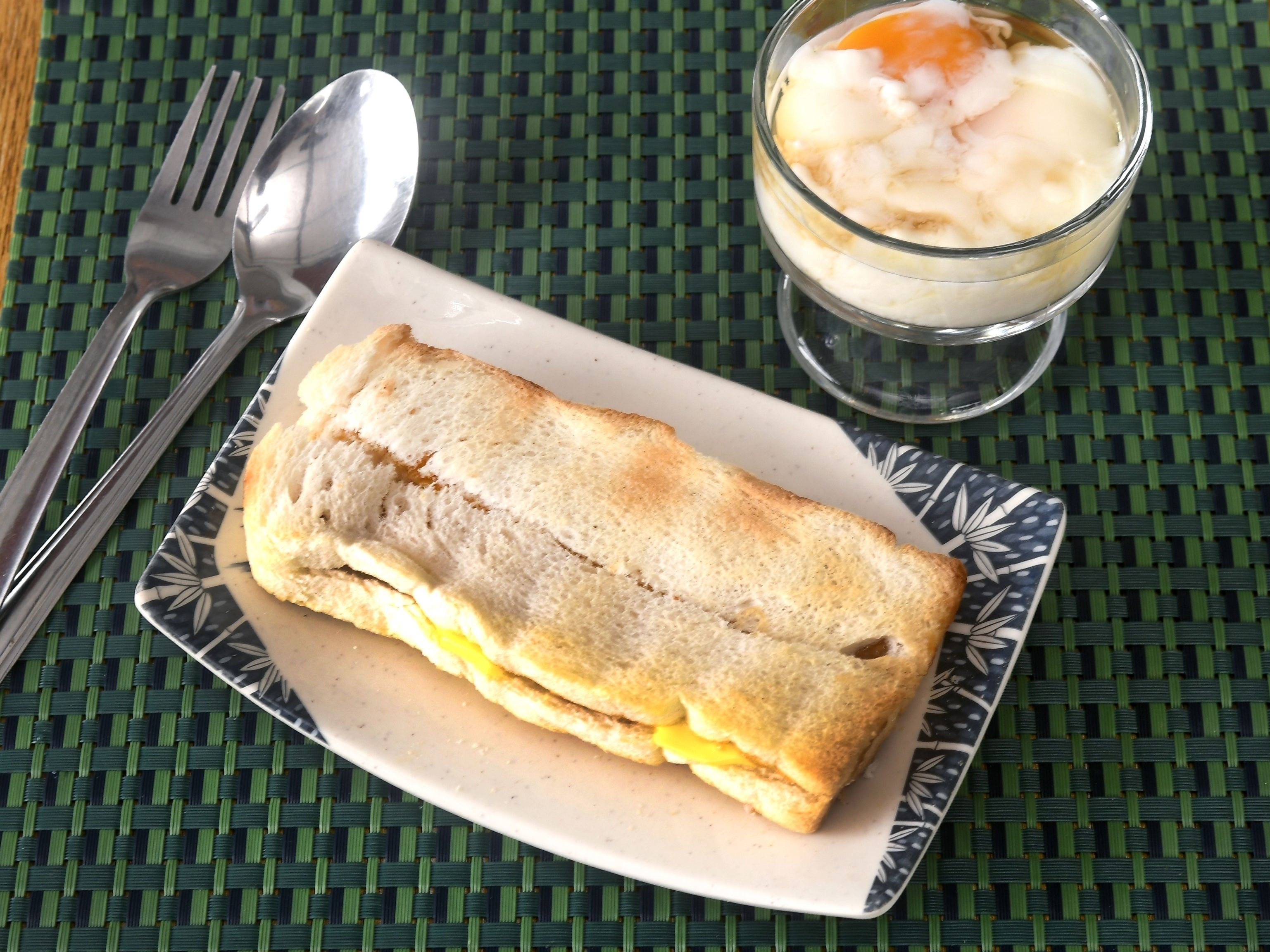 A sandwich on a plate with a spoon and fork beside it, and a glass of dessert on a woven mat.