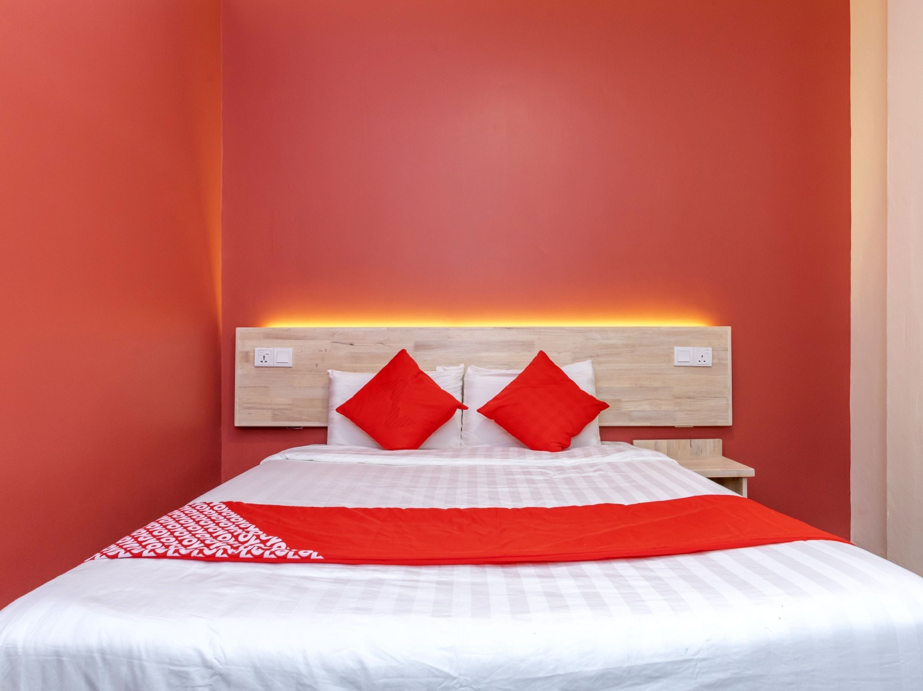 A hotel room with a bed, red pillows, a red throw blanket, and a wooden headboard with a lighted panel.