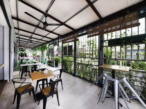 An indoor cafe with tables and chairs, large windows with plants, and a ceiling fan. - OYO 778 BMW 2 Hotel (Muar)