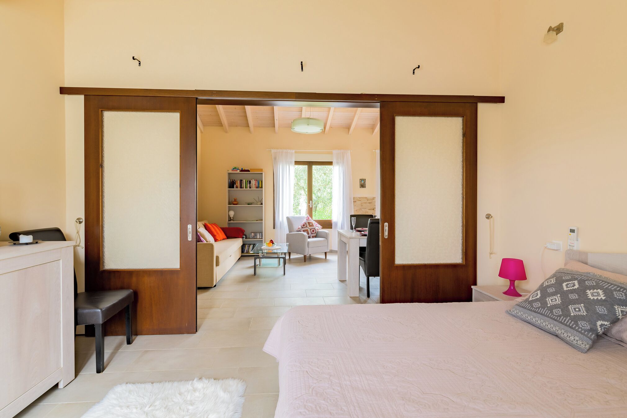 Een ruime slaapkamer met een groot bed, een bureau en een stoel. Achtergrond toont een boekenkast en een raam met gordijnen.