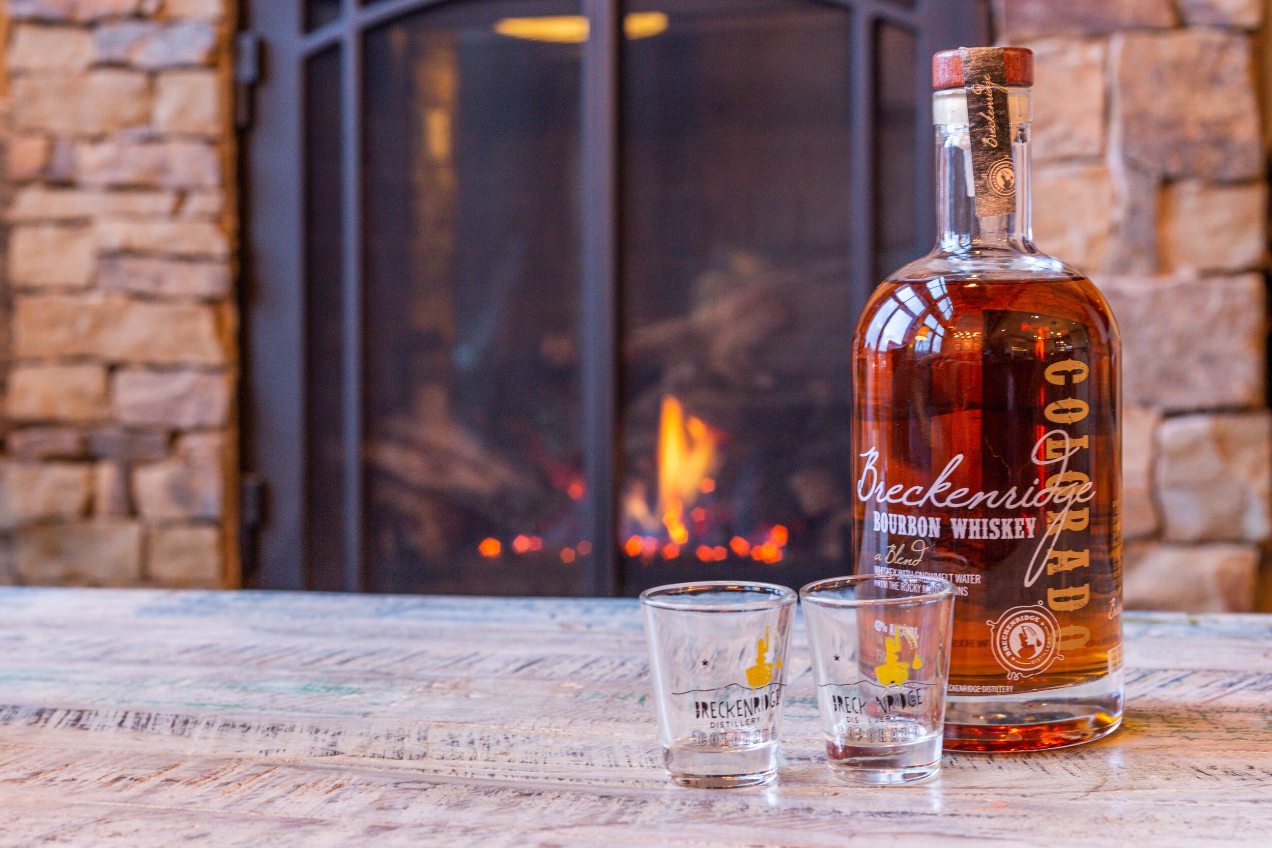 Eine Flasche Breckenridge Bourbon Whiskey und zwei Glasröhren auf einem Holztisch, im Hintergrund eine Ziegelwand und ein Kamin.
