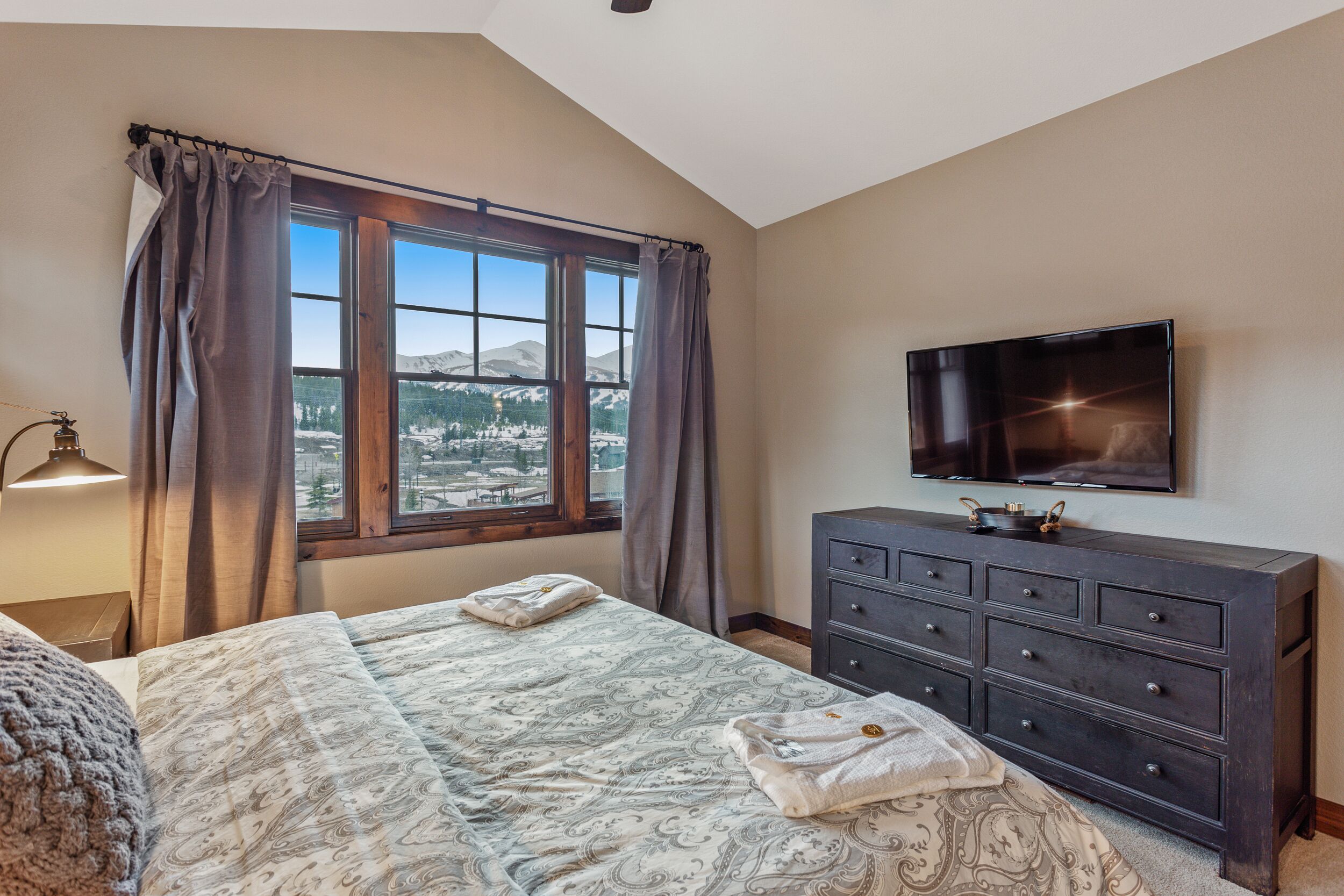 A bedroom with a large bed, a dresser, a television, and a window with a view of mountains.