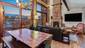 Un vaste espace de vie doté d’une grande table en bois, de sièges confortables, d’une cheminée et d’une grande fenêtre offrant une vue sur les montagnes.