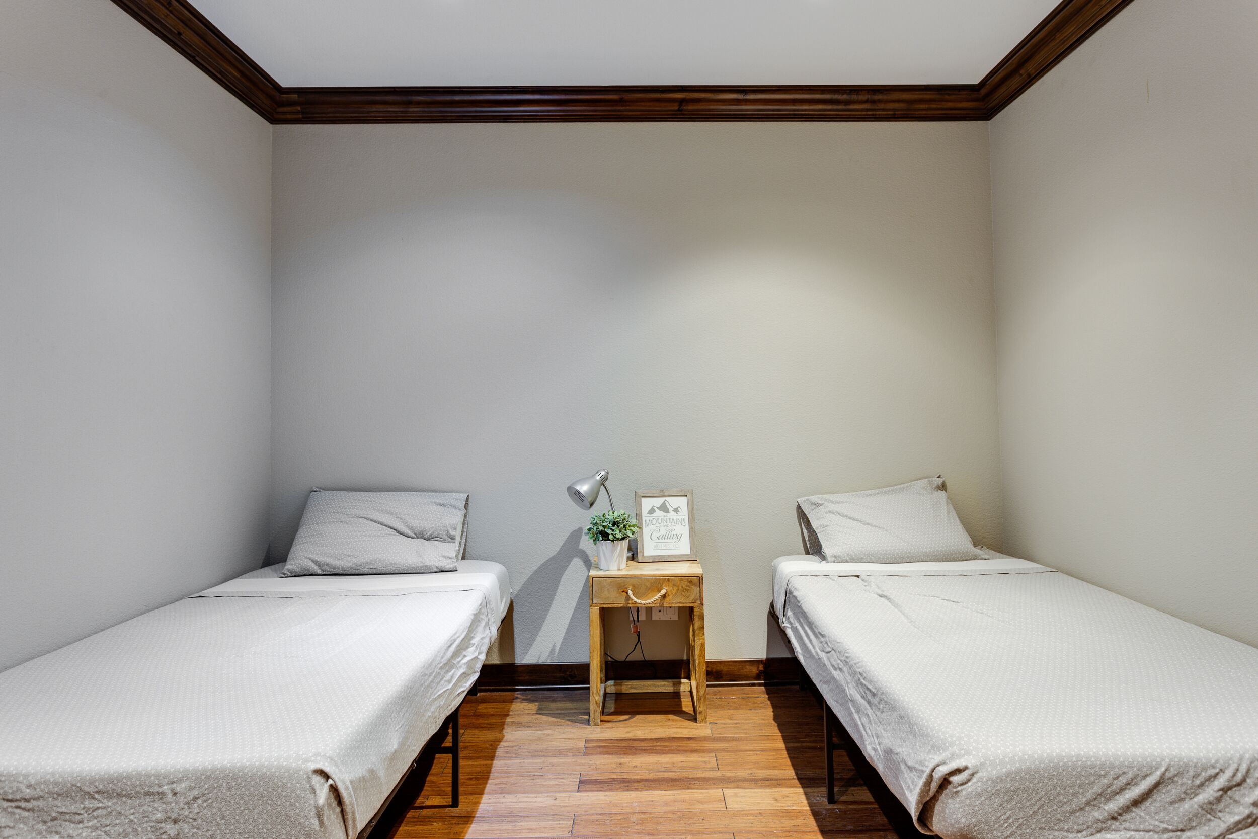 Ein kleines, sauberes Zimmer mit zwei Einzelbetten, einem Nachttisch aus Holz mit Lampe und einer kleinen Pflanze, sowie einem gerahmten Bild an der Wand.