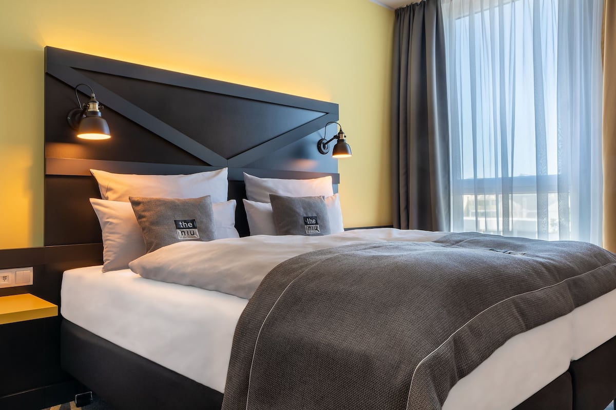 A modern hotel room with a large bed, a headboard with integrated lighting, and a view of the outdoors through sheer curtains.