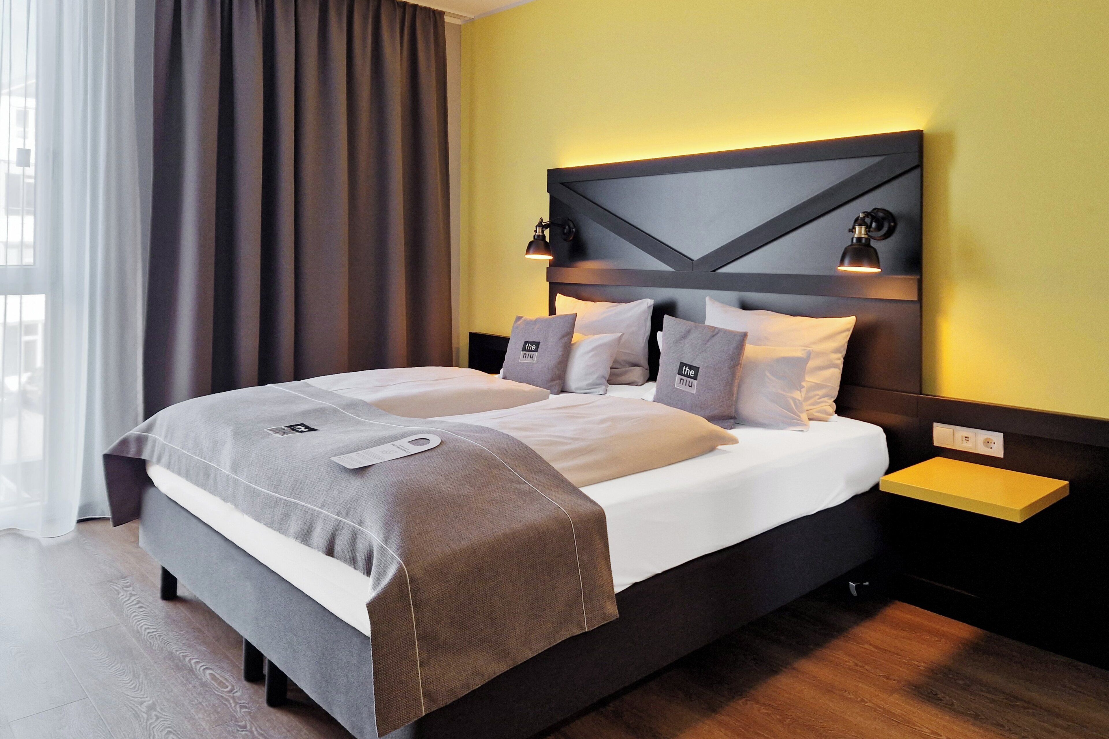 A modern hotel room with a large bed, a bedside table, a nightstand, and a window with curtains.