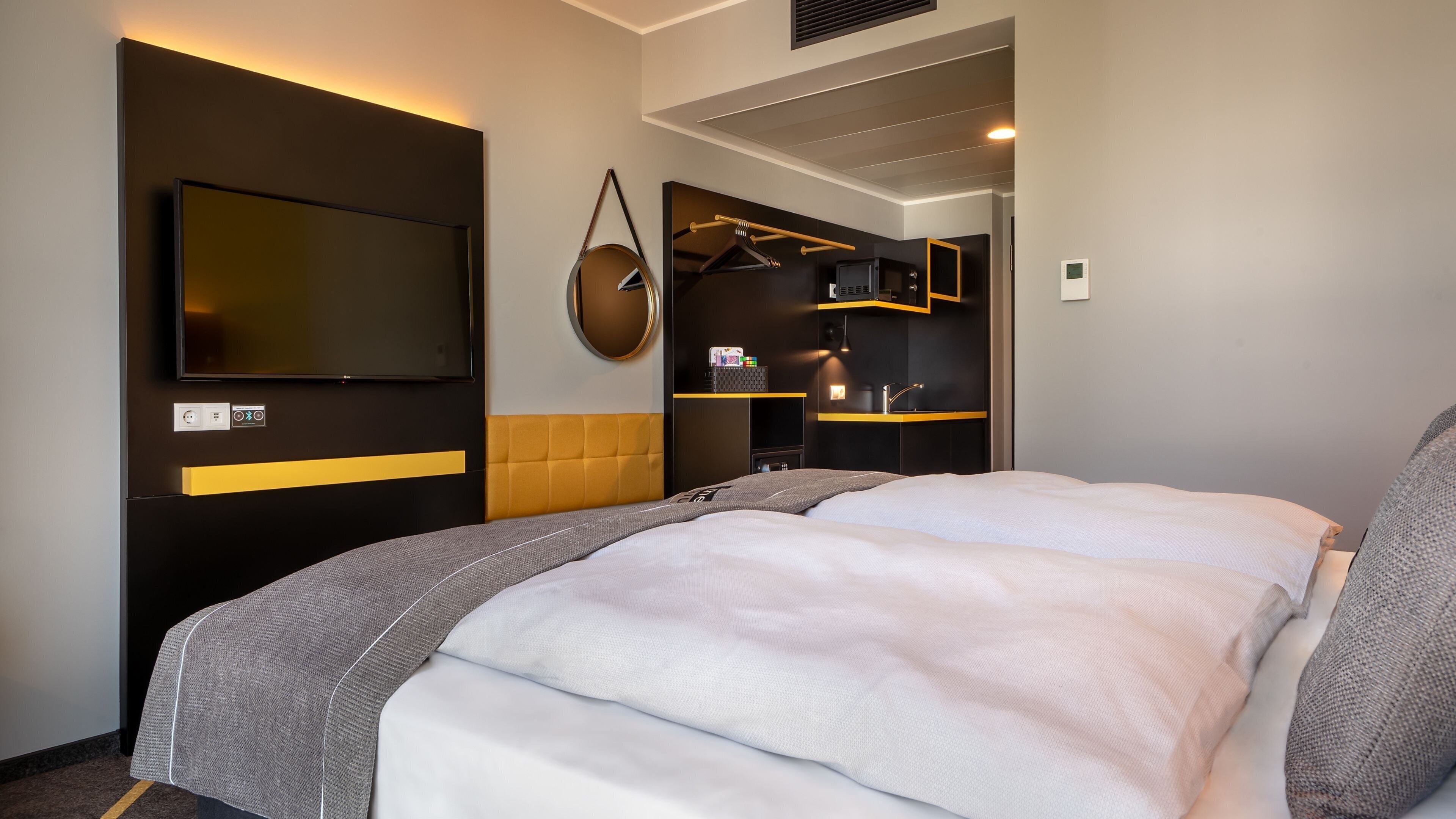 A modern hotel room with a large bed, a flat-screen TV, a wardrobe, and a small kitchenette.