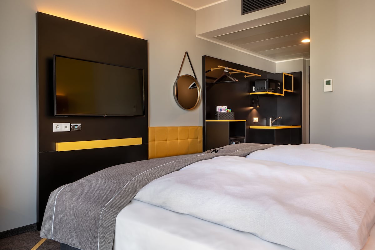 A modern hotel room with a large bed, a flat-screen TV, a wardrobe, and a small kitchenette.
