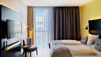 Quarto de hotel com cama, escrivaninha, cadeira, televisão e janela com cortinas.