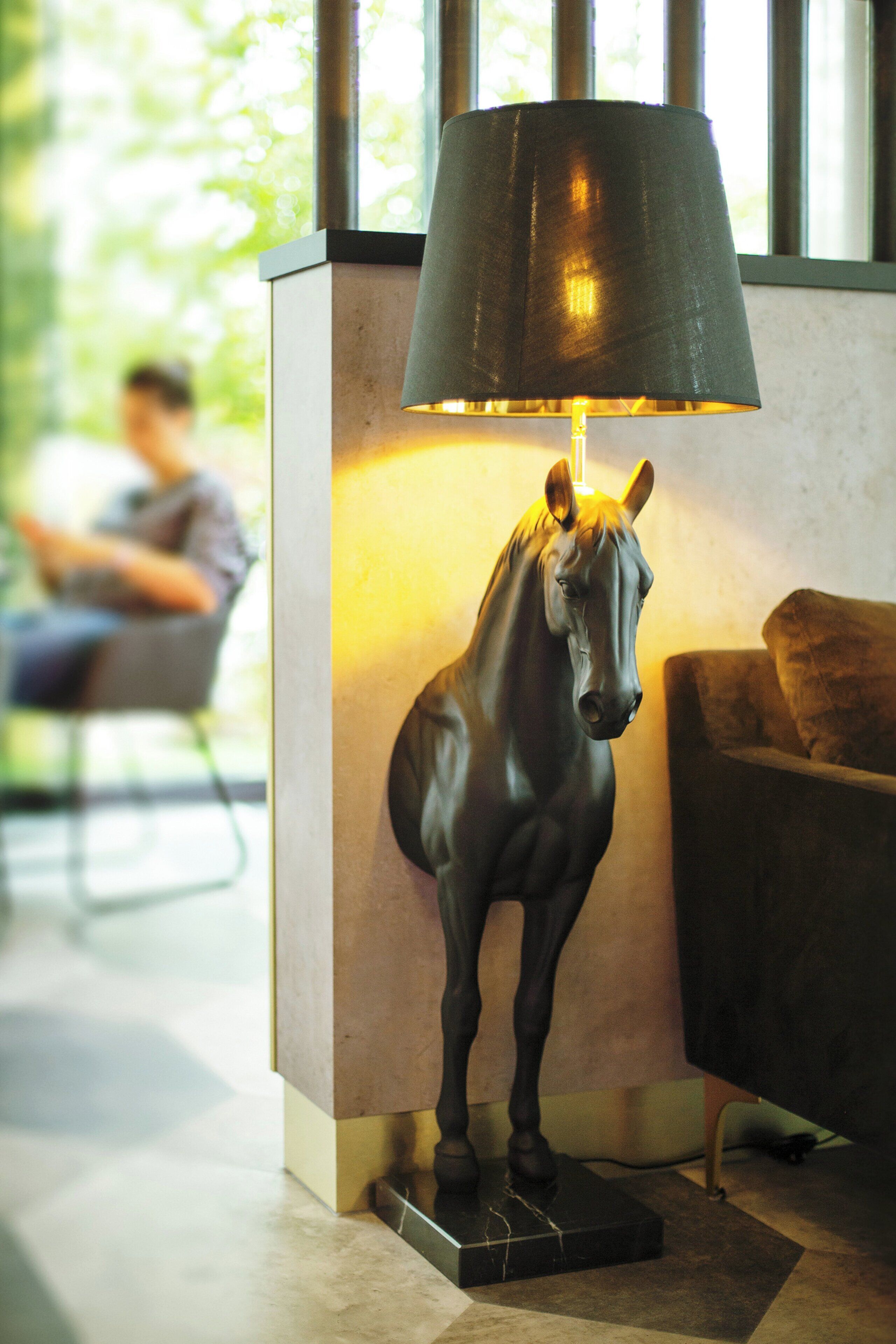 A statue of a horse on a pedestal with a lamp above it, placed in a room with a couch and a window in the background.