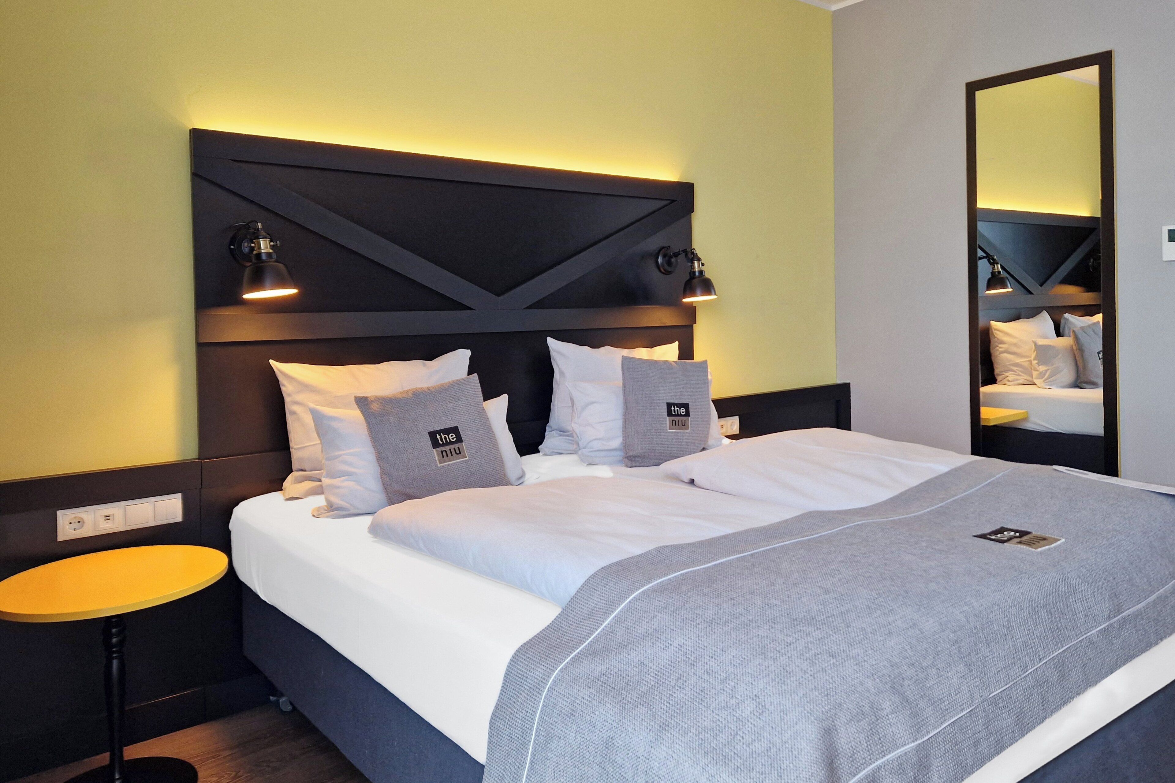 Habitación de hotel con una cama grande, una mesita de noche, un espejo y una lámpara fijada a la pared.