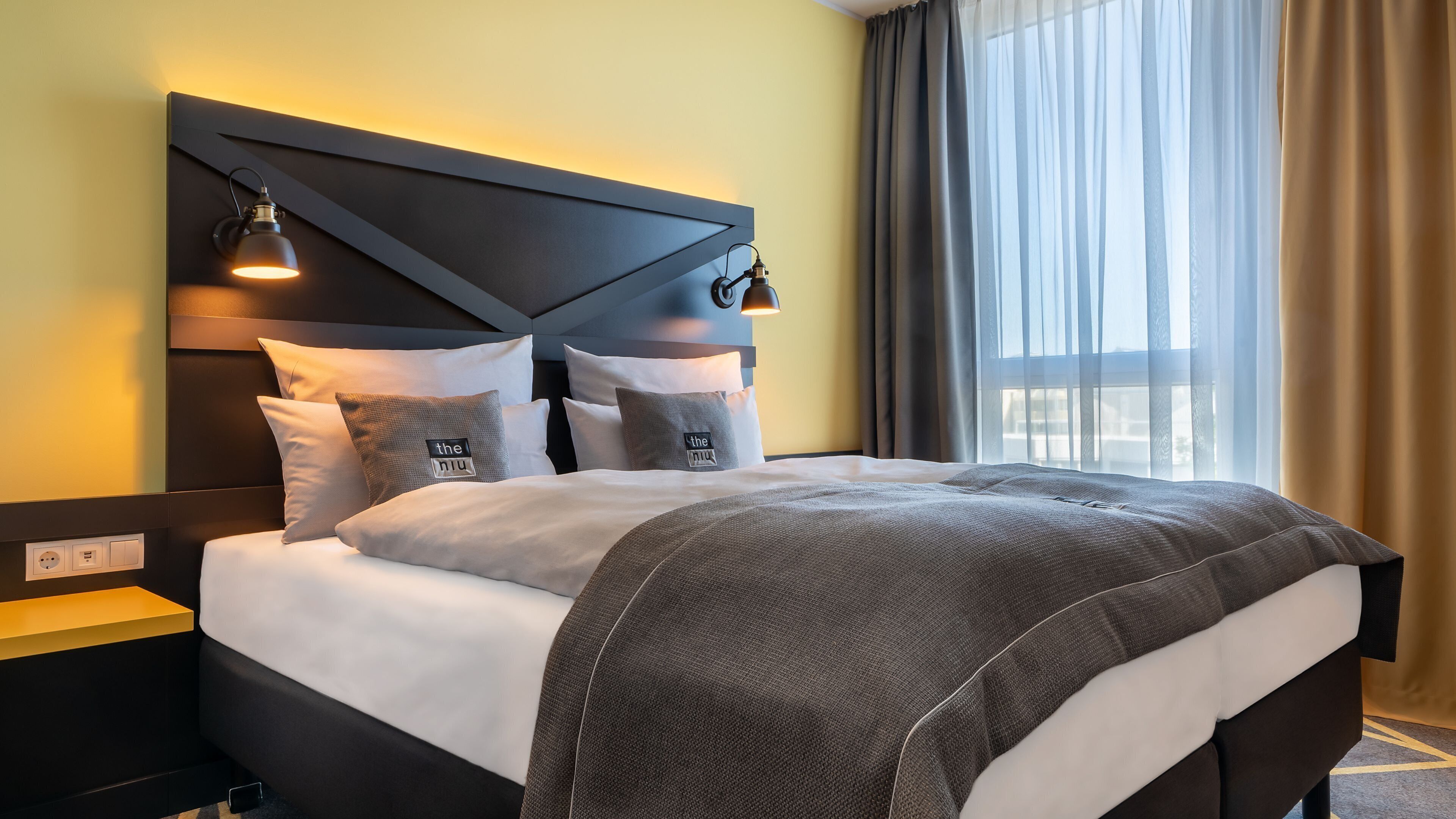 A modern hotel room with a large bed, a headboard with integrated lighting, and a view of the outdoors through sheer curtains.