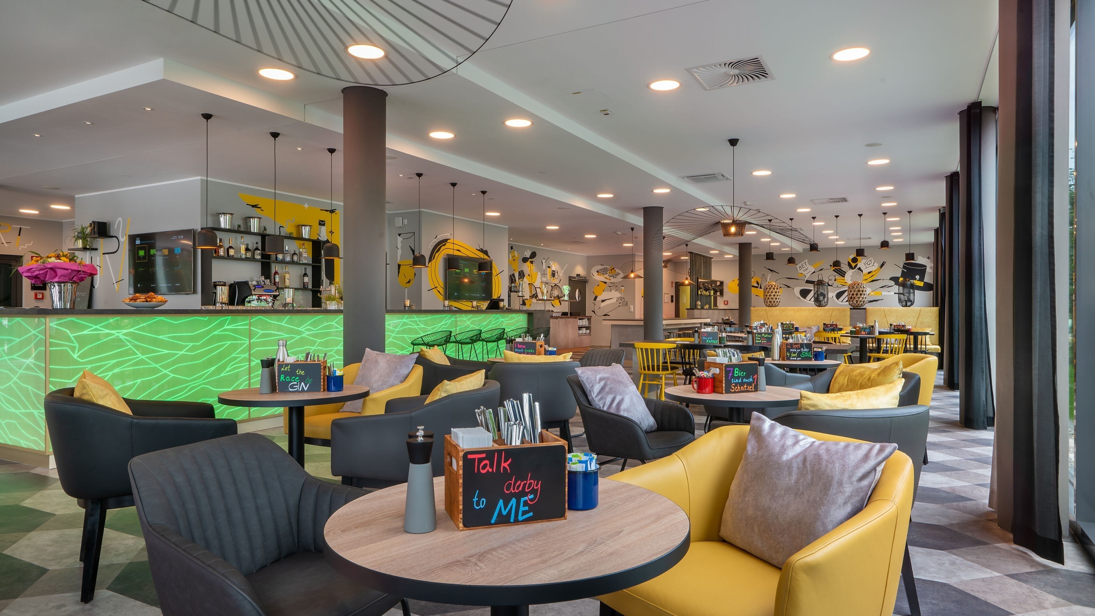 En modern bardel med sittplatser, en bardisk med en grön, bakbelyst skärm och en vägg med en gul och svart, abstrakt design.