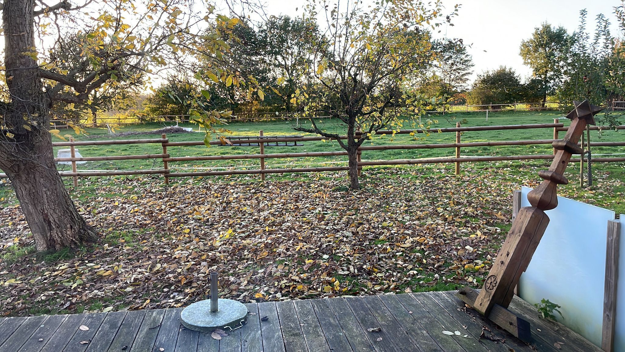 A wooden deck with a metal stand, a fence, and trees with yellow leaves in the background.