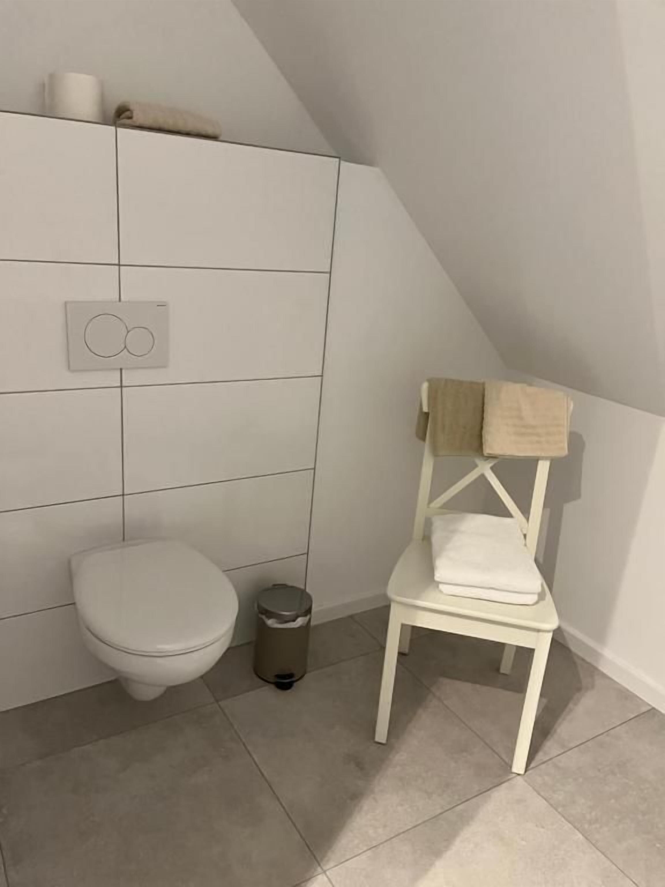 A bathroom with a toilet, a trash bin, and a white chair with a towel on it.