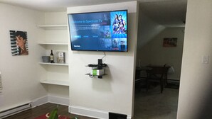 Un salon avec un téléviseur à écran plat fixé au mur, une petite étagère en dessous et une plante au premier plan.