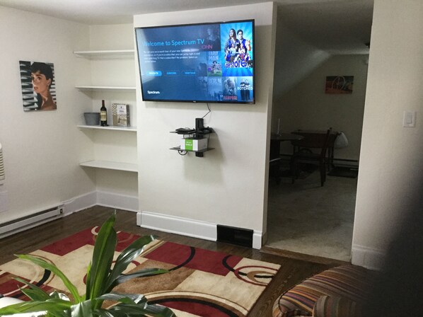 A living room with a flat-screen TV mounted on the wall, a shelf with decorative items, a plant, and a patterned rug.