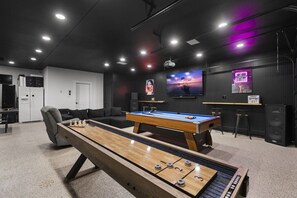 A billiards table, a pool table, a sofa, and a television in a room with dark walls and ceiling lights.