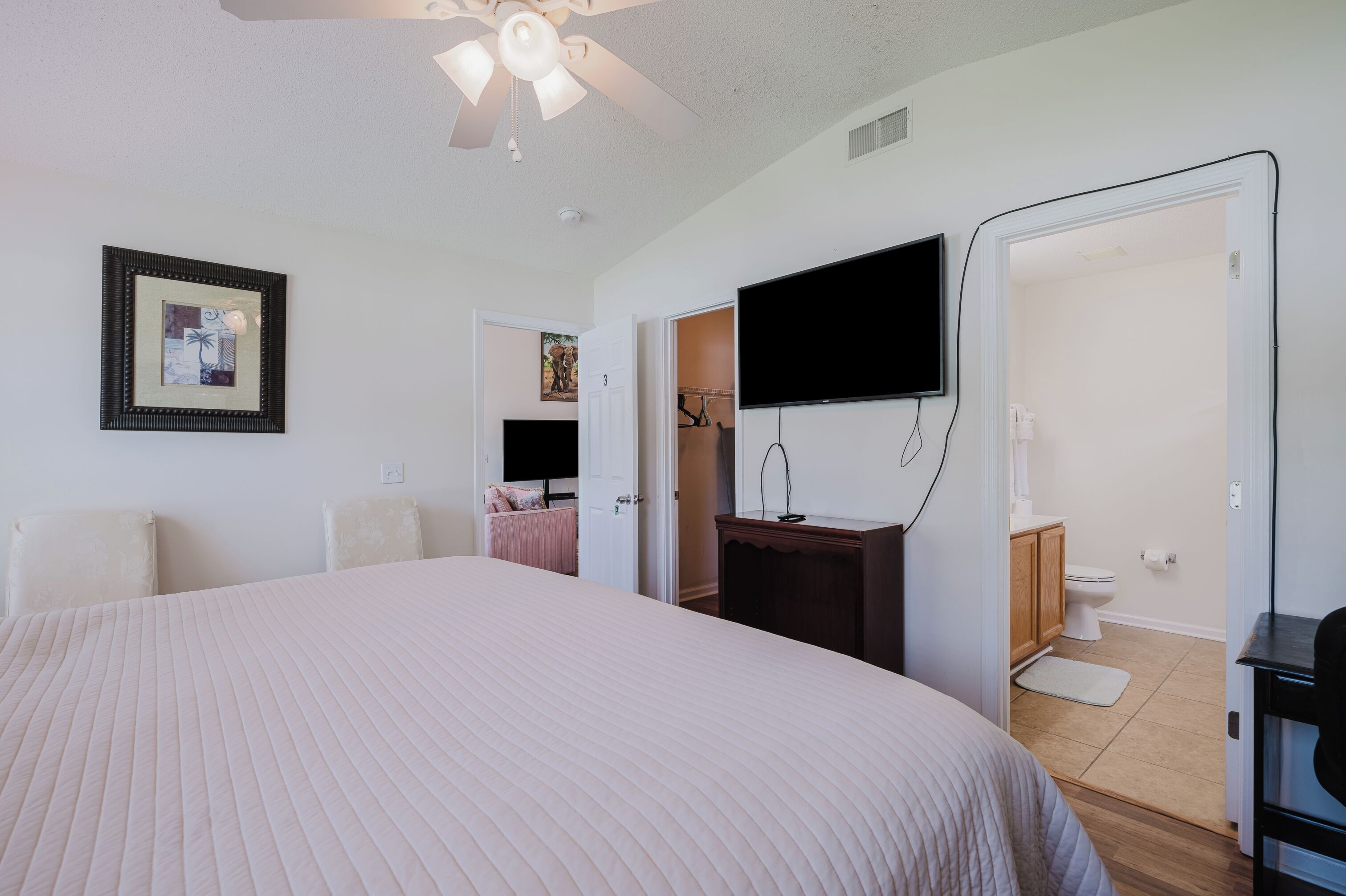 A bedroom with a large bed, a ceiling fan, a TV mounted on the wall, and a door leading to a bathroom.