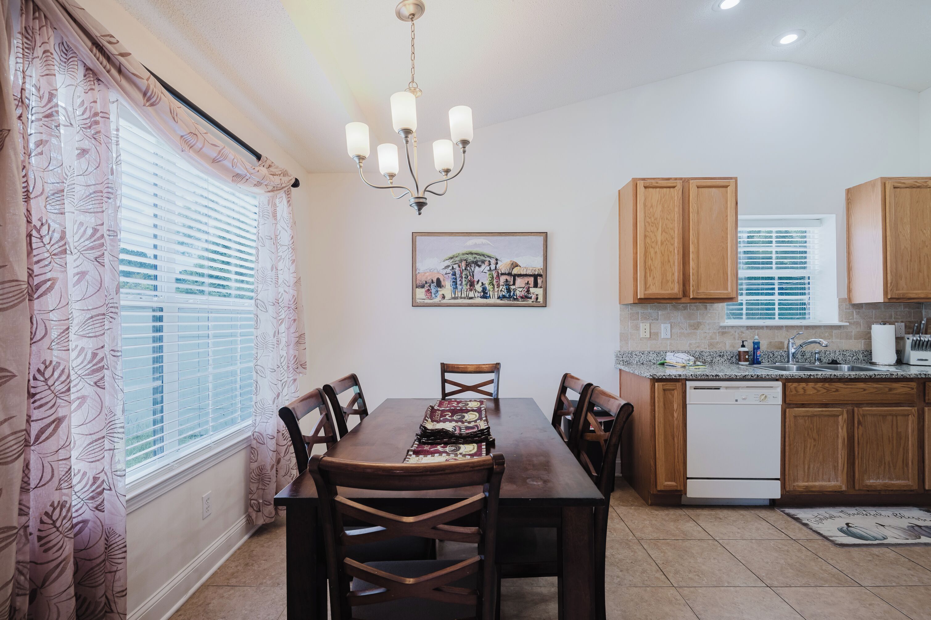 A dining area with a wooden table and chairs, a chandelier, and a kitchen with wooden cabinets and a dishwasher.