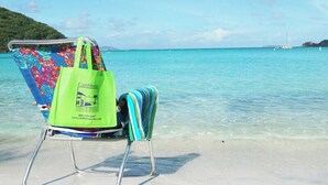 Ein Liegestuhl mit buntem Blumenmuster und einer grünen Tasche an einem Sandstrand, im Hintergrund klares türkisblaues Wasser und ein Segelboot.
