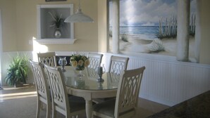 Et spiseområde med et rundt glasbord, hvide stole og et murmaleri med et strandmotiv.