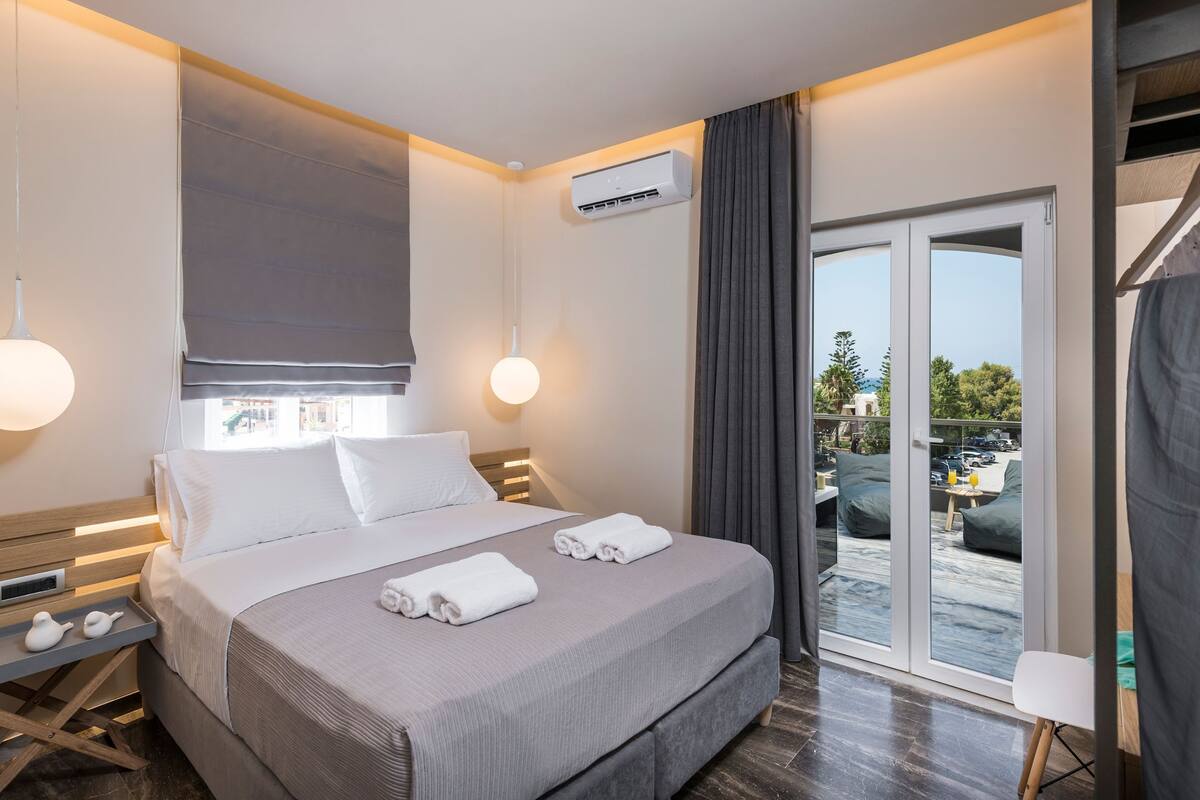A modern hotel room with a large bed, a bedside table with a lamp, a window with curtains, and a view of the outside.