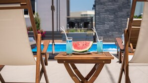 A poolside setting with wooden lounge chairs and a table with a watermelon and drinks. - Sun Ray Luxury Apartments (Chania)