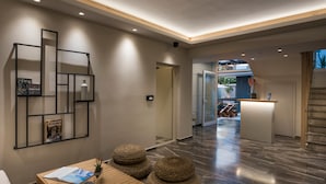 A modern hotel lobby with a reception desk, a seating area with a wooden table and poufs, and a geometric wall art installation. - Sun Ray Luxury Apartments (Chania)