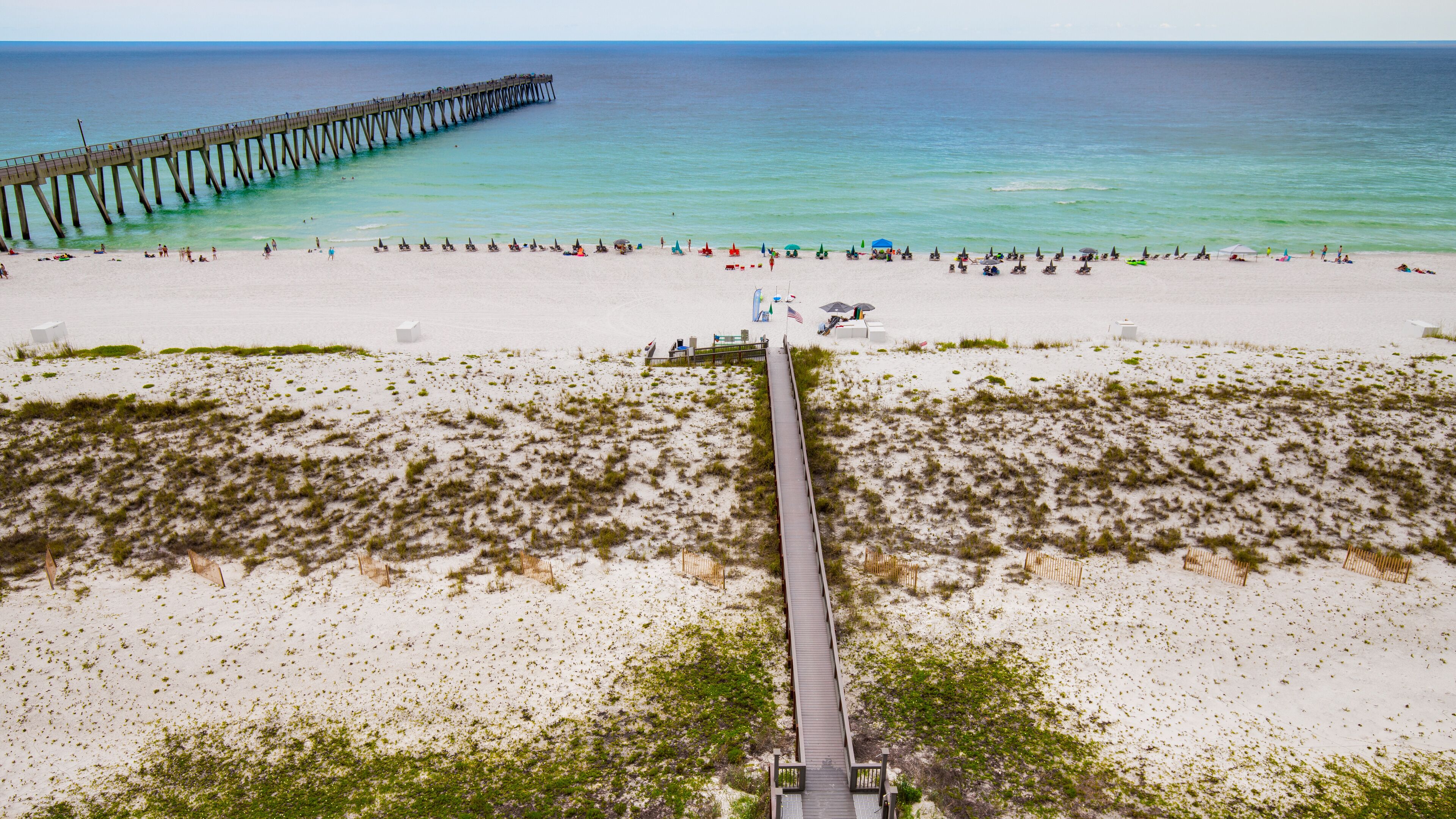 A beach with a wooden boardwalk leading to the water, a pier extending into the sea, and beachgoers enjoying the area.