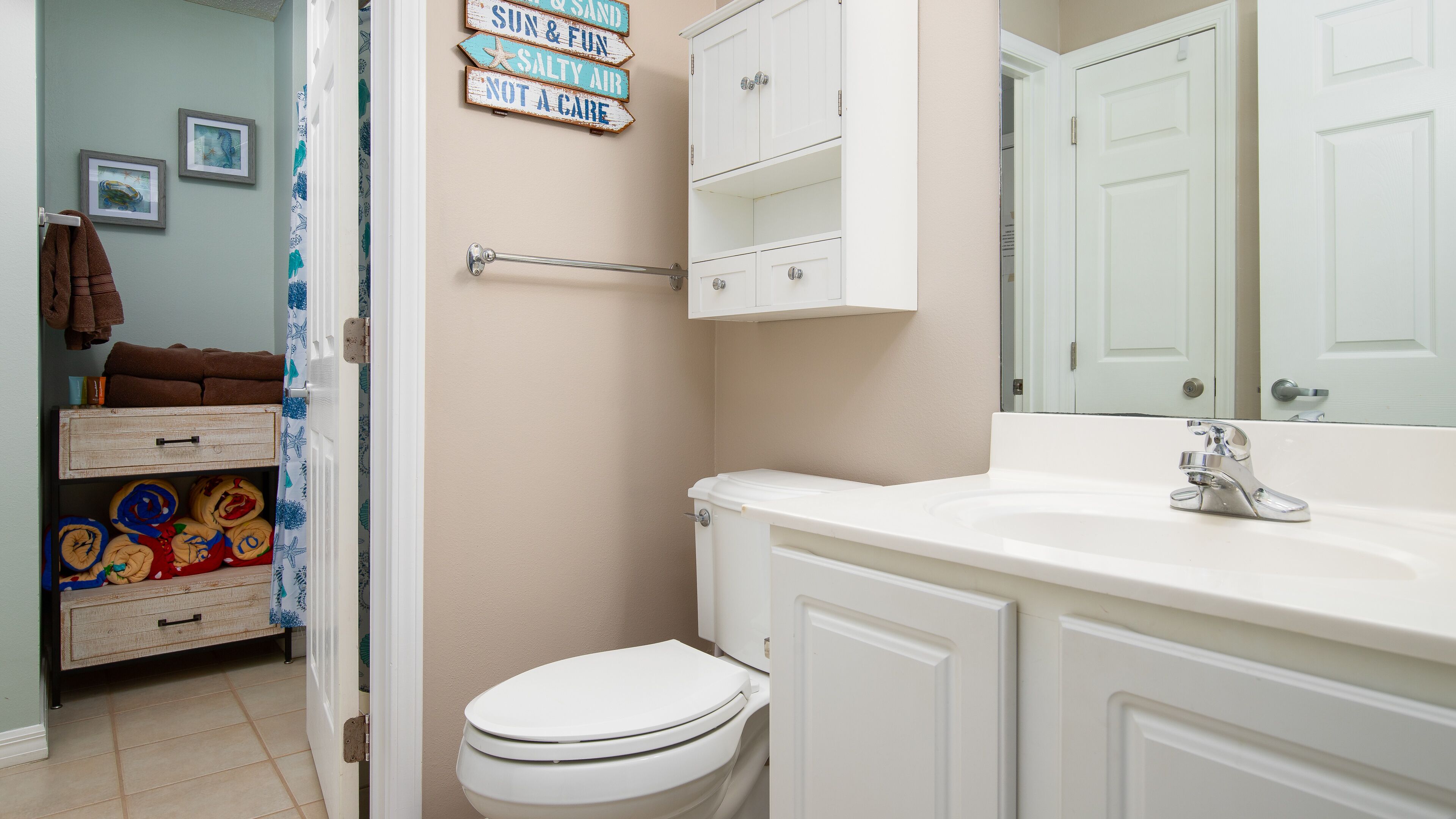 A bathroom with a white toilet, a sink with a chrome faucet, and a cabinet above the sink. There are towels stacked on a shelf and a sign on the wall.