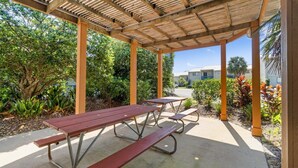 Une terrasse extérieure couverte, dotée de tables de pique-nique, entourée de verdure et avec un bâtiment en arrière-plan.