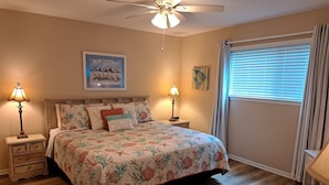 Une chambre à coucher comprenant un lit, deux tables de chevet, un ventilateur de plafond, une fenêtre avec des stores et des œuvres d’art accrochées au mur.