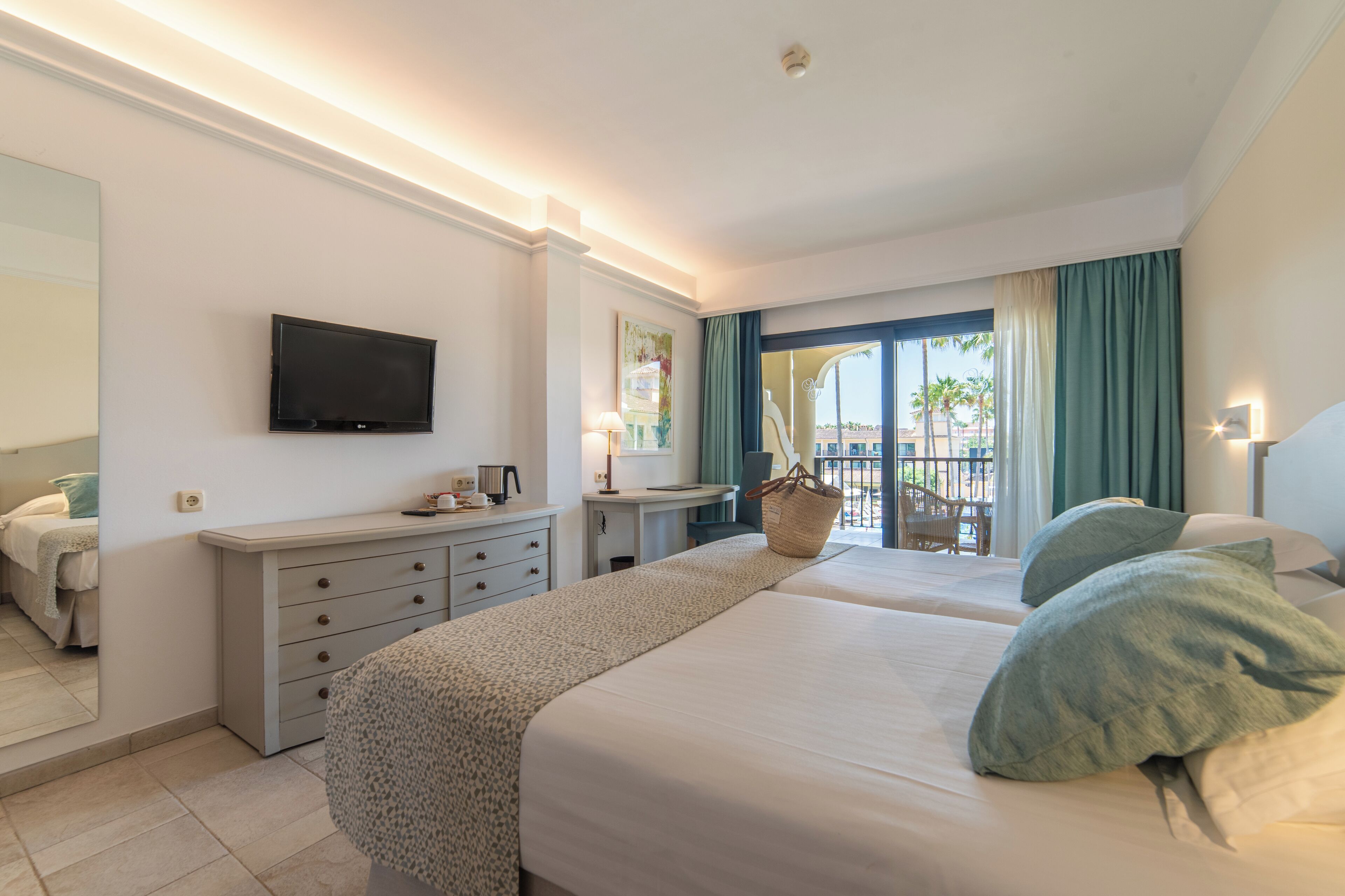 Habitación de hotel con dos camas, televisión, escritorio y balcón con vistas a palmeras.