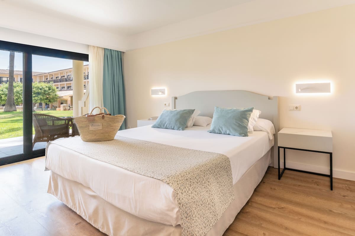 A hotel room with a large bed, a bedside table, a nightstand, a window with curtains, and a view of the building outside.