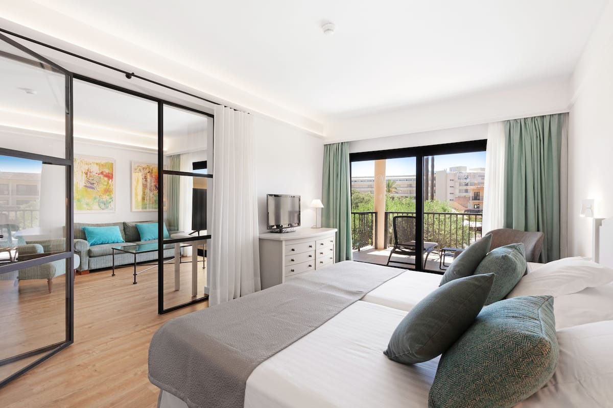A modern hotel room with a large bed, a TV, a balcony with a view of the city, and a dining area with a sofa and chairs.