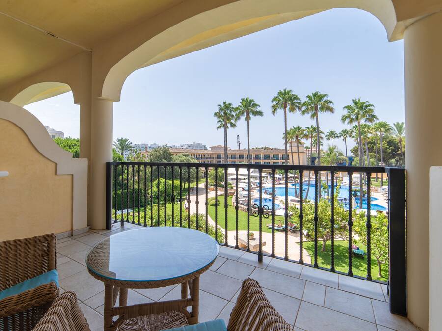 Un balcone con mobili in vimini, un tavolino in vetro e vista su una piscina e palme.
