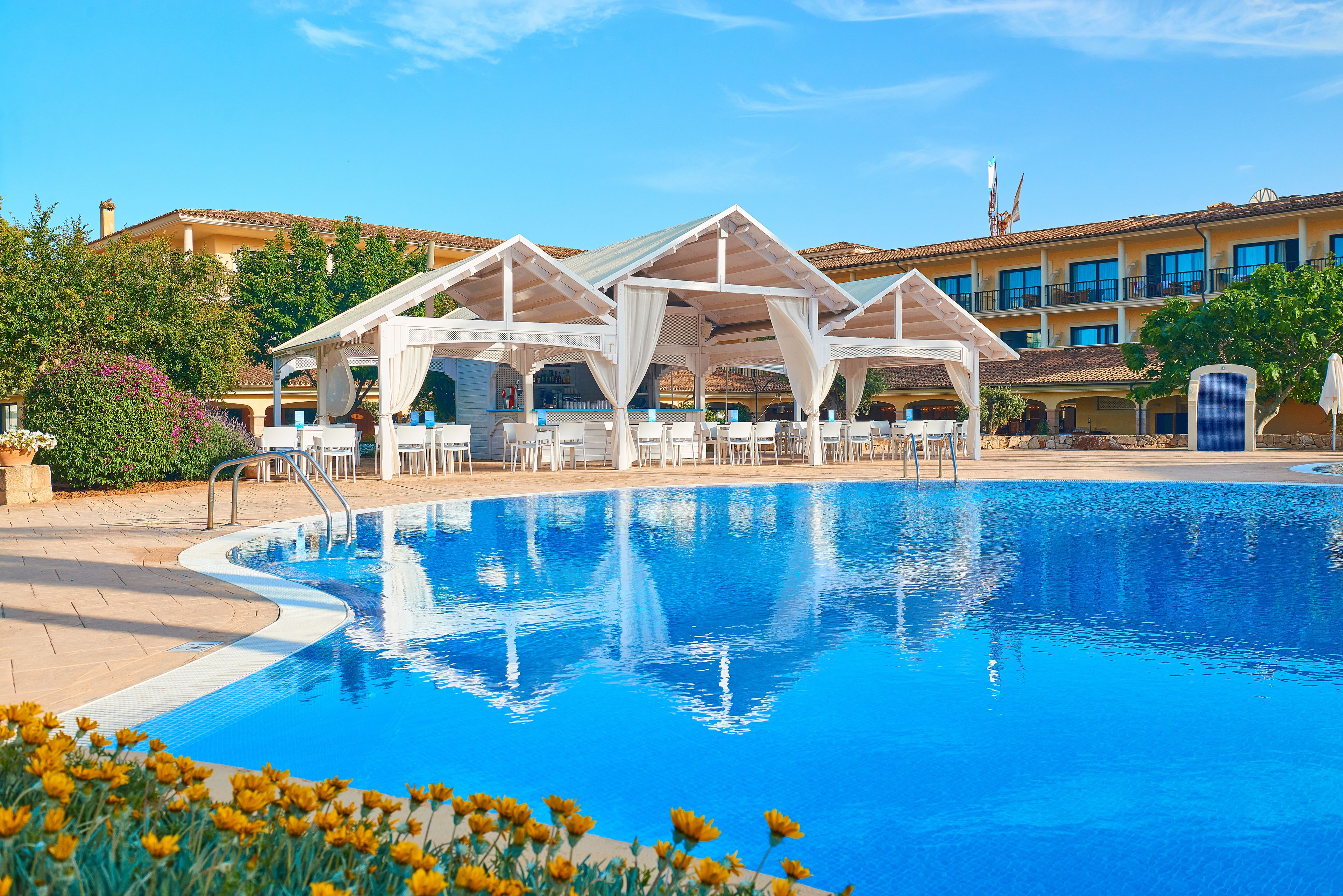 Una piscina con toldos blancos y sillones de descanso, rodeada por un edificio con varios balcones y un cielo azul despejado.
