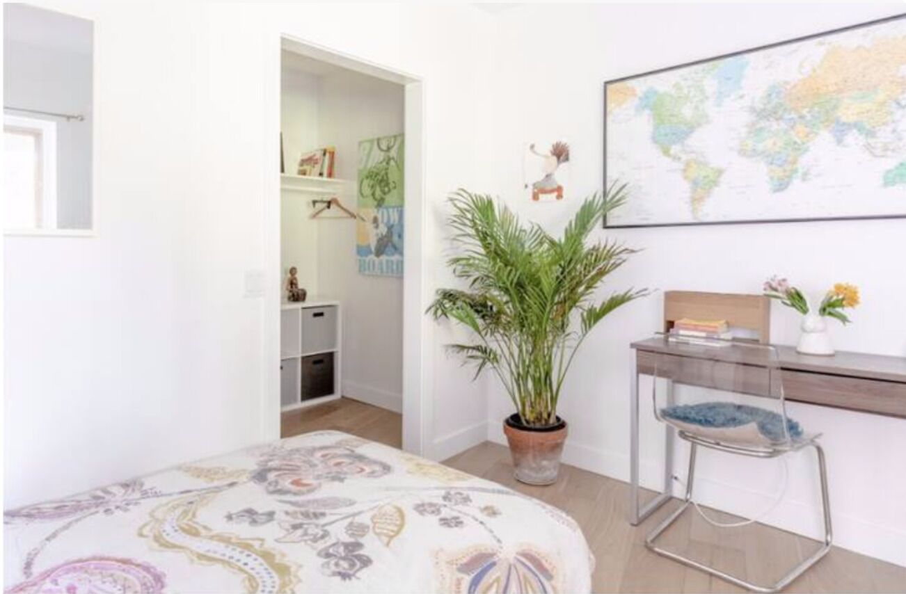 A bedroom with a bed, a desk with a chair, a potted plant, a world map on the wall, and a door leading to another room.