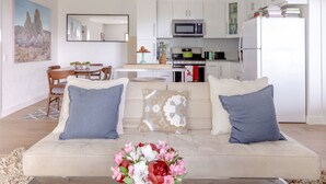Una sala moderna con un sofá beige, dos cojines azules y un centro de mesa floral. Se aprecia un comedor con una mesa y sillas de madera al fondo.