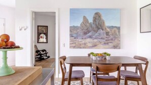 Un comedor con una mesa lista para cuatro personas, un cuadro de un paisaje desértico y un frutero verde.